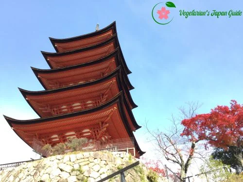 Five-story red pagoda on a stone base against a clear blue sky, with autumn trees nearby.