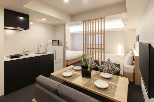 Open-plan studio with kitchenette, a four-seat dining table, gray sofa, and a bed separated by a wooden slat divider.